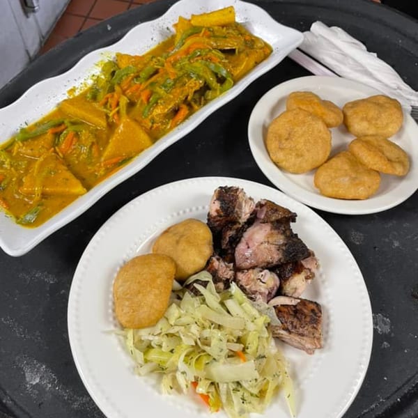 Plates of Jamaican curry, jerk chicken, fried dumplings, and cabbage on a dark surface.