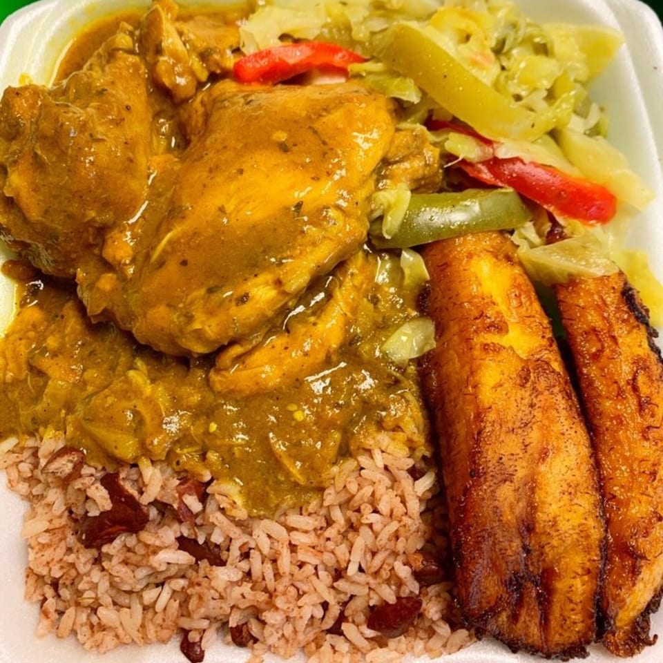 Curry chicken with rice and peas, fried plantains, and steamed cabbage in a takeout container.