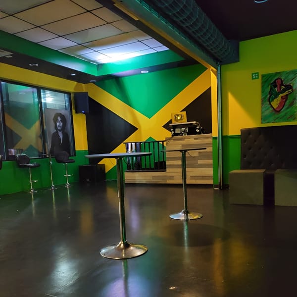 A vibrant Jamaican-themed bar interior with green and yellow walls, high-top tables, and a DJ booth.