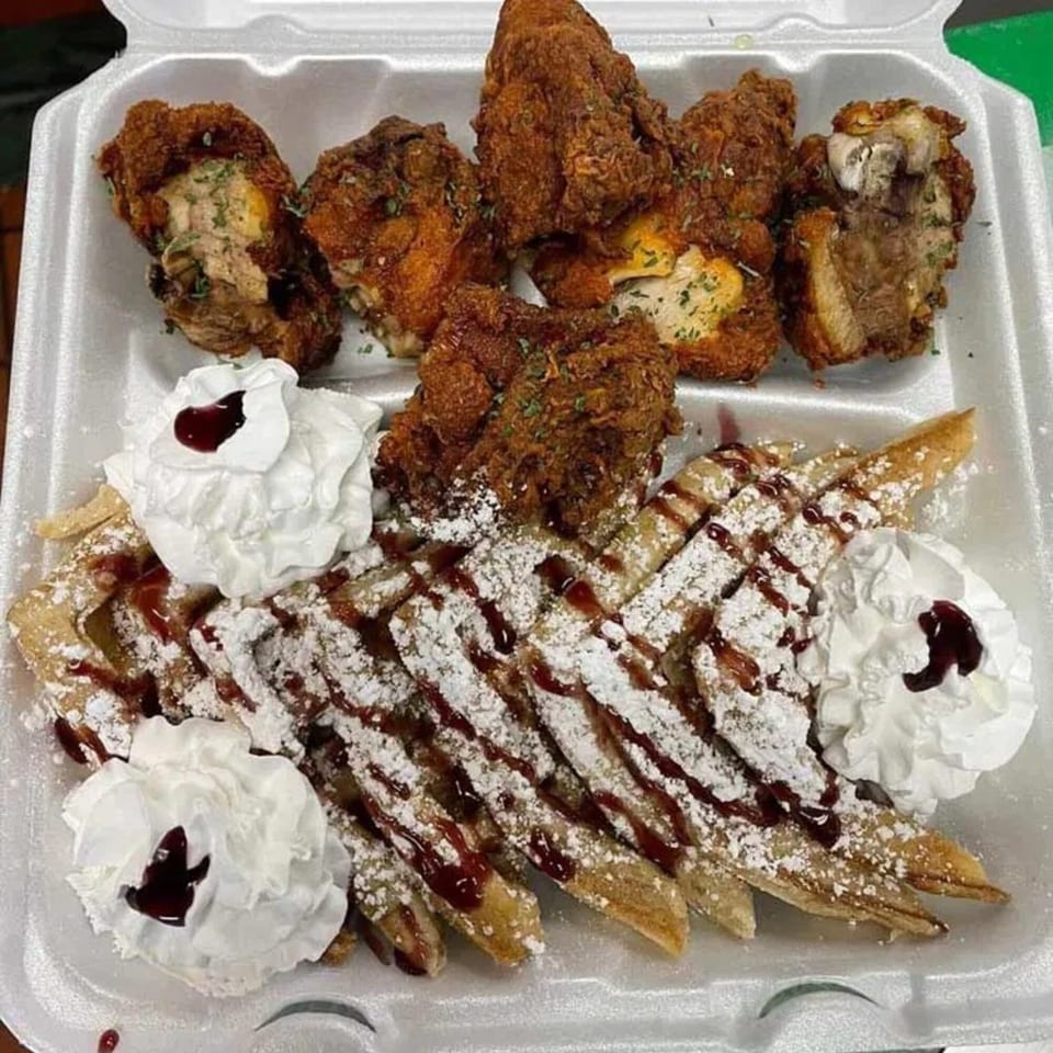 Fried chicken and waffles with powdered sugar, chocolate drizzle, and whipped cream in a takeout container.