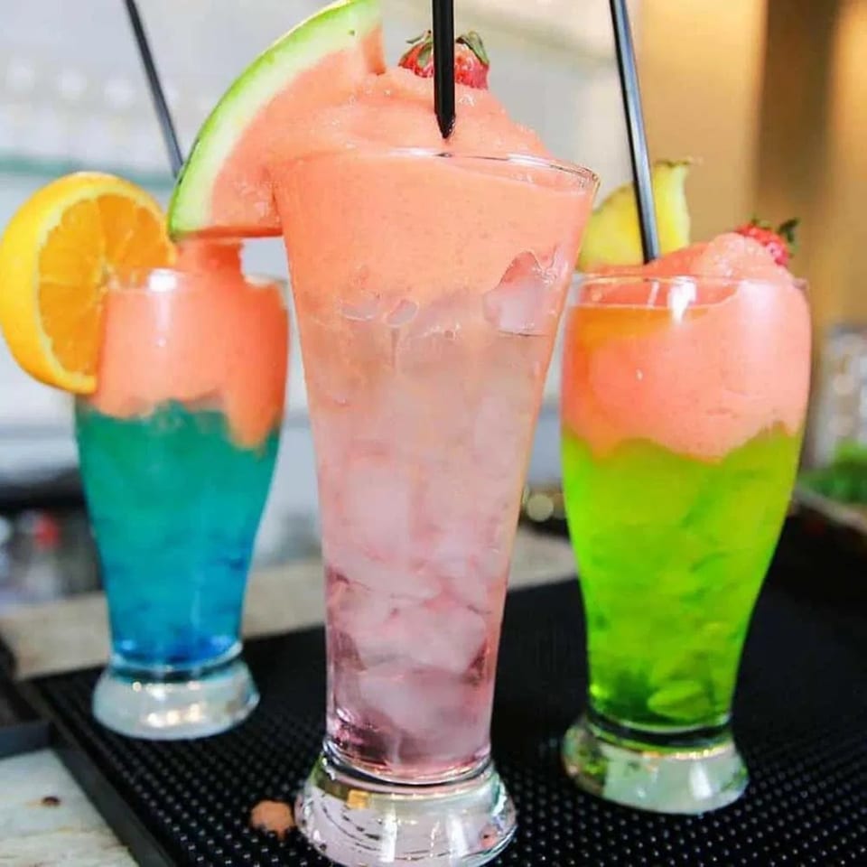 Three vibrant, layered frozen drinks with fruit garnishes, including watermelon, orange, and strawberry.