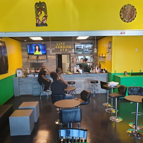 Interior of Litt Jamaica 876 restaurant with a bar, patrons, and high-top tables.
